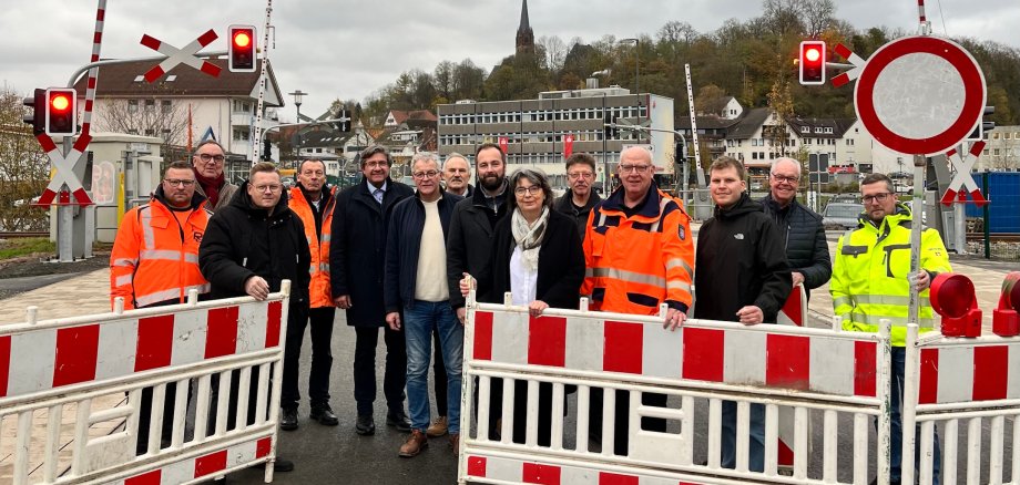 Menschengruppe hinter einer Straßenabsperrung, dahinter ein Bahnübergang